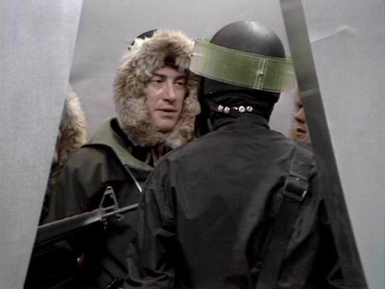 Claude 3.7 describes the image as: "This image appears to be from a science fiction television series from the late 1970s or early 1980s. It shows a tense confrontation scene in what looks like a sterile, institutional corridor or doorway. 

On the left is a person wearing a fur-trimmed parka hood, facing off against what appears to be a security guard or soldier in dark clothing wearing a helmet with a visor. The guard is holding what looks like a weapon.

The scene has the distinctive visual …
