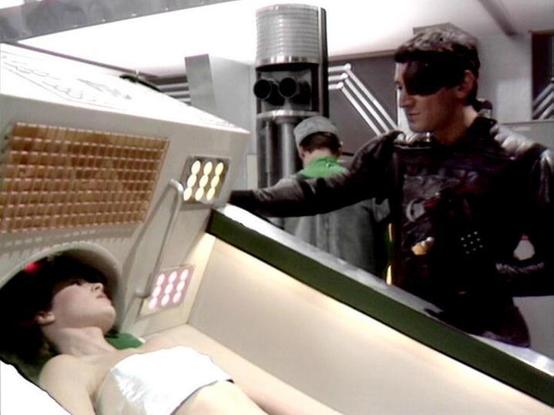Claude 3.7 describes the image as: "This image appears to be from a science fiction television series, showing a futuristic medical or examination scene. In the foreground, there's a person lying on what looks like a medical bed or examination table, wearing what appears to be a white garment. Standing nearby is another individual dressed in dark clothing with some sort of eye covering or visor, reaching toward a control panel with illuminated sections. The setting has a distinctly retrofuturis…