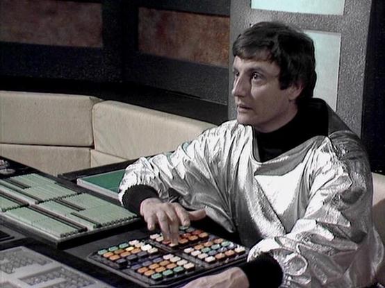 Claude 3.7 describes the image as: "This image appears to be from a science fiction television series from the late 1970s or early 1980s. It shows a person in a futuristic silver/metallic jacket with a black turtleneck underneath, seated at what looks like a control console or computer terminal. 

The console has numerous colorful buttons and controls, suggesting a spacecraft or advanced control room setting. The background shows what appears to be a modernist set design with cream-colored furn…