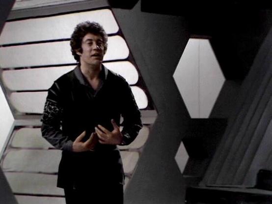 Claude 3.7 describes the image as: "The image shows a person in a dark outfit standing in what appears to be a futuristic corridor or spacecraft interior. They're wearing a black garment with some detailed embroidery or pattern on the sleeve, and appear to be in the middle of a conversation or dramatic moment.

The setting features distinctive architectural elements typical of science fiction productions from the late 1970s/early 1980s, with geometric white wall panels and hexagonal design elem…