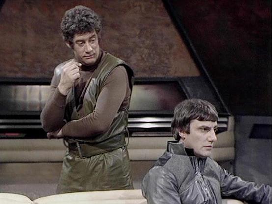 Claude 3.7 describes the image as: "This image shows a scene from a classic science fiction television series, set in what appears to be a spacecraft or control room with angular walls and a futuristic design typical of late 1970s/early 1980s British sci-fi production.

Two performers are pictured: one standing on the left wearing a olive-green/brown uniform with a vest-like outer layer, with curly hair and a contemplative expression; and another seated on the right wearing a gray high-collared…