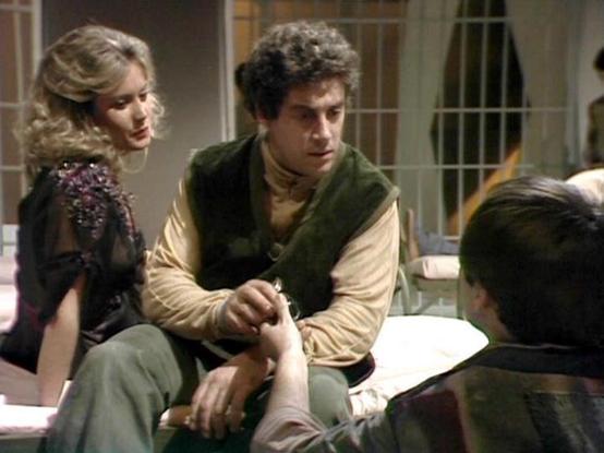 Claude 3.7 describes the image as: "This image appears to be from a vintage science fiction television series from the late 1970s or early 1980s. The scene shows three people in what looks like a spacecraft or futuristic interior setting. 

On the left is a person with blonde curly hair wearing an elaborate dark outfit with embellished details. In the center is someone in a light-colored shirt with a green vest or wrap. This character appears to be engaged in conversation or interaction with an…