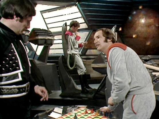 Claude 3.7 describes the image as: "The image shows a scene from a retro science fiction television series set on what appears to be a spaceship bridge or control room. Three people are visible in the frame - one wearing a black outfit with decorative elements on the left, another in a light gray jumpsuit with red trim on the right, and a third figure standing in the background. The futuristic set features control panels, unusual lighting, and what looks like a game board or control surface wit…