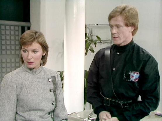 Claude Sonnet 4.0 describes the image as: "I can see this is a scene from what appears to be a science fiction television series, showing two people in what looks like a futuristic or clinical setting. The woman on the left is wearing a light-colored, military or uniform-style jacket with buttons, while the man on the right is dressed in a dark colored outfit. The setting appears to be some kind of medical facility or spaceship interior, with white walls and what looks like medical or technical…