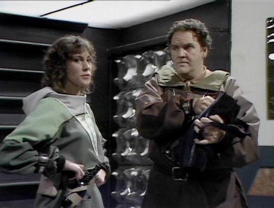 Claude 3.7 describes the image as: "This image appears to be from a science fiction television series, showing two individuals in a futuristic setting. They're wearing distinctive costume pieces that have a utilitarian, space-age aesthetic with mixed earth tones and metallic elements. The scene takes place in what looks like a spacecraft or space station interior with characteristic panel walls and technical elements visible in the background.

Both figures are equipped with what appear to be w…