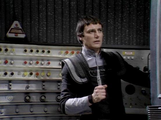Claude 3.7 describes the image as: "The image shows someone in a futuristic costume with a high collar and distinctive black and silver/gray design. They appear to be in what looks like a control room or technical area with numerous buttons, lights, and control panels visible in the background. The setting has a classic science fiction aesthetic with its utilitarian control panels featuring colored buttons and indicators.

The costume design is characteristic of vintage sci-fi television produc…