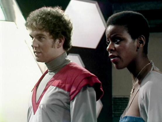 Claude Sonnet 4.0 describes the image as: "I can see two people in what appears to be a futuristic spacecraft or space station setting. One person has curly hair and is wearing a distinctive red and white uniform with gray accents that has a somewhat military or official appearance. The other person is wearing what looks like a light-colored outfit. The setting has the characteristic sterile, technological aesthetic typical of science fiction productions from this era, with white and geometric …