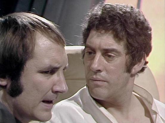 Claude 3.7 describes the image as: "The image shows two characters in what appears to be a sci-fi television production from the late 1970s or early 1980s. The setting looks like the interior of a spacecraft or futuristic facility with light-colored walls visible in the background.

Two men are engaged in conversation, with one on the left having shorter, lighter hair and sideburns, while the person on the right has curly, darker hair. They're wearing light-colored clothing that appears to be d…