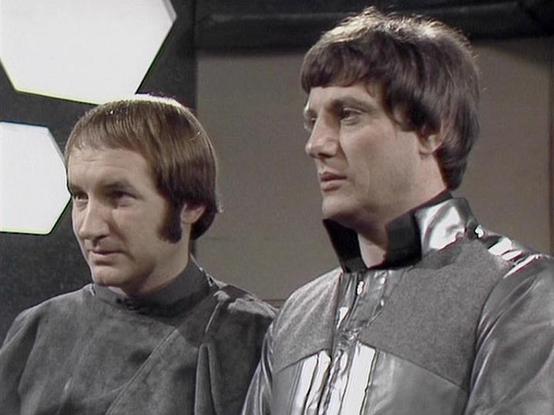 Claude 3.7 describes the image as: "The image shows two people in a science fiction setting, wearing futuristic costumes with metallic and gray elements typical of classic sci-fi television production design from the late 1970s/early 1980s. 

The individuals are dressed in similar styled uniforms with high collars, though the person on the right has more prominent silver detailing on their costume. Both have distinctive period hairstyles that align with the aesthetic of British television produ…