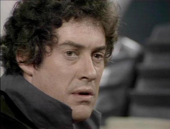 Claude 3.7 describes the image as: "The image shows a person with curly dark hair and an intense expression, appearing to be in a dramatic scene. They're wearing what looks like a dark coat or jacket with a high collar, typical of science fiction costume design from the late 1970s or early 1980s.

The shot is a close-up with a blurred background, capturing a moment of apparent tension or concern. The lighting and film quality have the distinctive look of British television productions from that…
