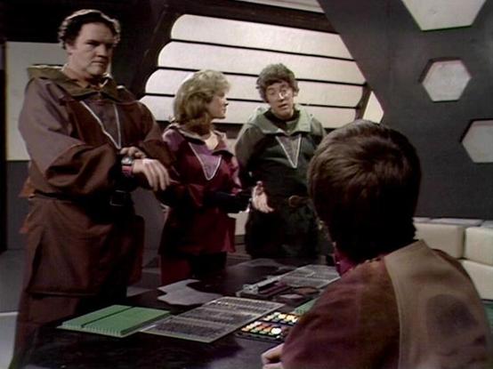 Claude Sonnet 4.0 describes the image as: "This scene takes place in what appears to be the flight deck or control room of a spacecraft, featuring the characteristic hexagonal wall panels and control consoles typical of the series' futuristic aesthetic. Four crew members are gathered around a central control station, with one person seated at the console operating equipment while three others stand nearby observing. The setting suggests this is during a mission briefing or tactical discussion, …
