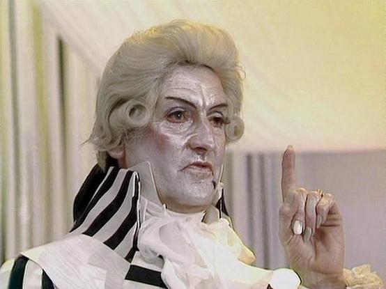 Claude Sonnet 4.0 describes the image as: "I can see this is a scene featuring an elaborately costumed character in period dress, including a powdered wig and ornate clothing with black and white striped elements. The character appears to be making a pointing gesture and has theatrical makeup or prosthetics that give them a distinctive, somewhat grotesque appearance. The setting appears to be an interior location with pale yellow/cream colored walls, suggesting an elegant or formal environment.…