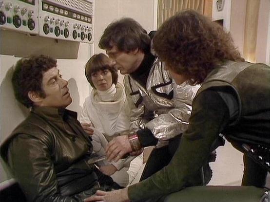 Claude 3.7 describes the image as: "This image appears to be from a vintage science fiction television series from the late 1970s or early 1980s. The scene shows a tense moment in what looks like a spacecraft or facility control room. 

There are four people in the image - one person wearing a dark leather jacket seated or leaning against equipment, another in a white outfit with a distinctive collar, and two others dressed in futuristic-looking costumes including what appears to be a silver/me…