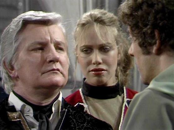 Claude 3.7 describes the image as: "The image shows a scene from what appears to be a science fiction television production from the late 1970s or early 1980s, based on the filming style and costume design. 

In the foreground are three people engaged in what seems to be a tense conversation. On the left is an older person with silver hair wearing formal attire with a distinctive collar. In the center is a person with blonde hair pulled back, wearing a red outfit with a black choker or high nec…