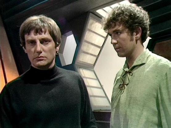 Claude Sonnet 4.0 describes the image as: "This scene appears to be set aboard a spacecraft, likely the Liberator, given the characteristic curved walls and technological interior design typical of the series. The lighting creates a dramatic atmosphere with shadows and metallic surfaces visible in the background.

Two men are present in what appears to be a tense or serious moment. One wears a distinctive black leather outfit with a high collar, while the other is dressed in lighter colored clo…