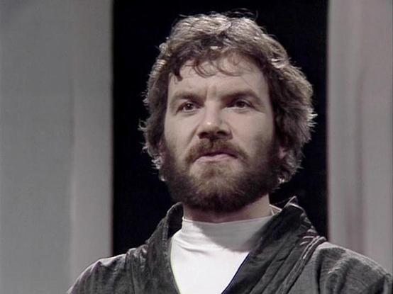 Claude Sonnet 4.0 describes the image as: "I can see this appears to be from a science fiction television production, likely from the late 1970s or early 1980s based on the visual style and production quality. The image shows a person with curly dark hair and a beard, wearing what appears to be a dark colored garment over a light colored shirt. The setting looks like it could be aboard a spacecraft or in a futuristic facility, with clean, sterile-looking walls or panels visible in the backgroun…
