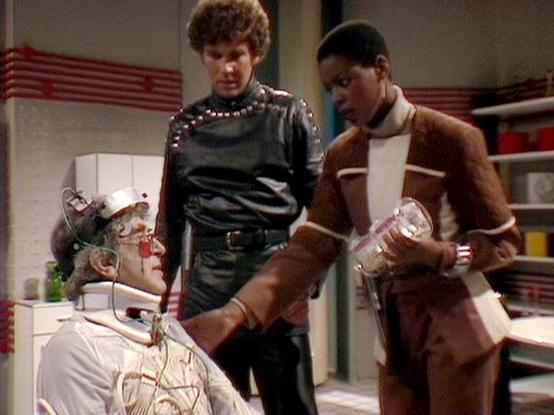 Claude 3.7 describes the image as: "This image appears to be from a science fiction television series, showing a scene set in what looks like a futuristic laboratory or medical facility. 

The scene features three people - one person seated wearing a white outfit with various wires or technological components attached to their head, possibly representing some kind of experiment or medical procedure. Standing nearby are two others - one in a black leather-like outfit with decorative studs or emb…