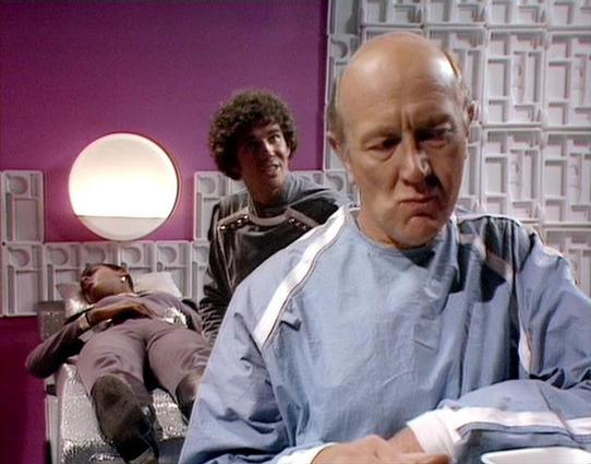 Claude Sonnet 4.0 describes the image as: "I can see this appears to be from a science fiction television series, showing what looks like a medical or scientific facility with futuristic set design. The scene features someone in medical/surgical attire in the foreground, with other figures visible in the background. The setting has distinctive sci-fi elements including curved walls, geometric patterns, and ambient lighting typical of 1970s-80s television production design. The scene appears to …