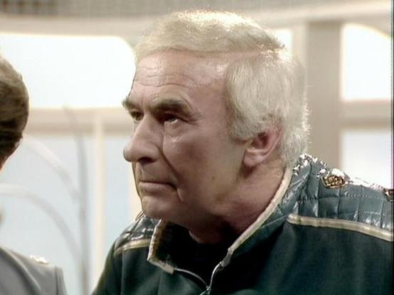 Claude 3.7 describes the image as: "The image appears to be from a classic British television production, likely from the late 1970s or early 1980s based on the visual quality and aesthetic. 

The scene shows an elderly person with white hair wearing a dark jacket with teal/green elements and decorative trim. The individual has a serious, contemplative expression and appears to be in a conversation within what looks like an interior setting with light-colored walls or panels visible in the back…