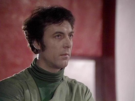 Claude 3.7 describes the image as: "The image shows a person with dark hair wearing a green turtleneck and what appears to be a light olive-colored jacket or vest. They have a serious, contemplative expression and are shown in profile against a reddish background. 

This appears to be a still from a science fiction television production from the late 1970s or early 1980s, based on the filming quality and styling. The costume design has that distinctive retro sci-fi aesthetic with simple, practi…
