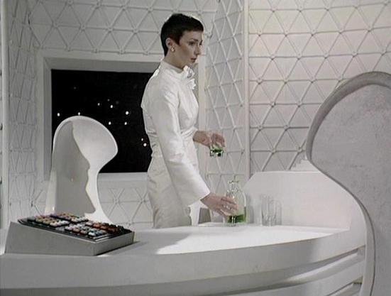 Claude 3.7 describes the image as: "The image shows a futuristic, minimalist white interior with a geometric patterned wall. A person with short dark hair is dressed in an elegant white outfit with a high collar, standing at what appears to be a curved white table or console. They're pouring a green liquid from a bottle into a glass, with additional glasses nearby. On the left side of the frame is what looks like a tray of colorful items, possibly controls or decorative elements. A stylish whit…