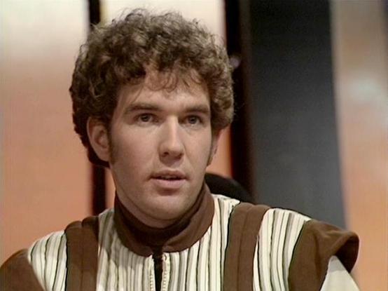 Claude 3.7 describes the image as: "The image shows a person with curly dark hair wearing a distinctive costume that features a cream/white striped garment with a brown vest or outer layer. The costume has a somewhat retro sci-fi appearance typical of British television productions from the late 1970s to early 1980s.

The setting appears to be a spaceship or futuristic interior with soft lighting and muted colors in the background. The production style has that characteristic look of television…