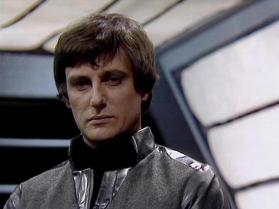 Claude Sonnet 4.0 describes the image as: "This image shows a character in a futuristic setting with sleek, metallic walls and angular architectural elements typical of science fiction spacecraft or space station interiors. The person is wearing a dark, high-collared uniform with a leather or synthetic material finish, consistent with the utilitarian yet stylish costume design of the series. The lighting creates dramatic shadows across their face, emphasizing the serious, contemplative expressi…