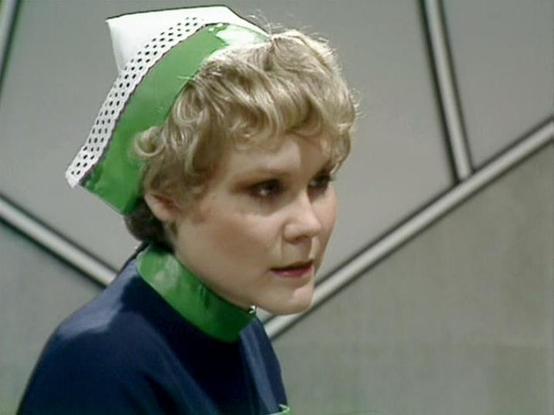 Claude 3.7 describes the image as: "The image shows a person wearing what appears to be a uniform with a distinctive green and white cap. They have light curly hair and are wearing a dark blue top with green accents around the collar. The background appears to be a futuristic or sci-fi setting with geometric wall panels.

This appears to be from a classic British science fiction television series from the late 1970s/early 1980s. The costume design is characteristic of that era's vision of futur…