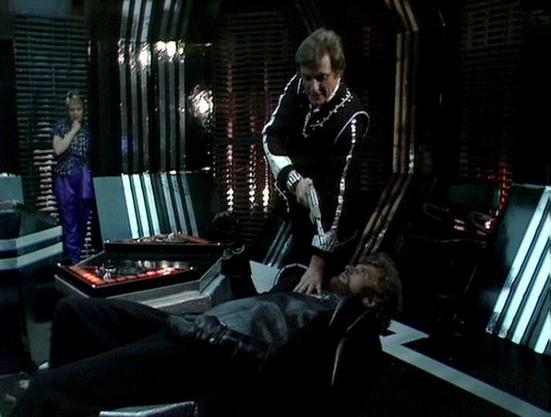 Claude Sonnet 4.0 describes the image as: "I can see this appears to be from a science fiction television series, showing what looks like a futuristic medical or technical facility. The scene shows someone lying on what appears to be an examination table or medical bed, with another person standing over them in dark clothing. There's a third figure visible in the background. The setting has the characteristic look of 1970s/80s sci-fi production design, with white and metallic surfaces, control …