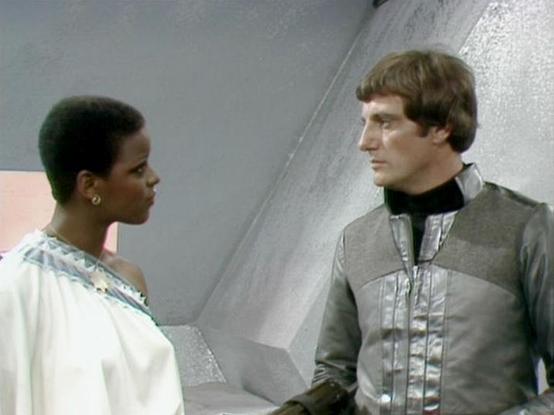 Claude Sonnet 4.0 describes the image as: "This appears to be a scene from a science fiction television series, showing two characters in what looks like a futuristic spacecraft or base interior. The setting features sleek, metallic walls with a distinctive angular design typical of sci-fi productions from that era.

One character is wearing an elegant white flowing garment with decorative trim, suggesting a formal or ceremonial outfit. The other character is dressed in what appears to be a mil…