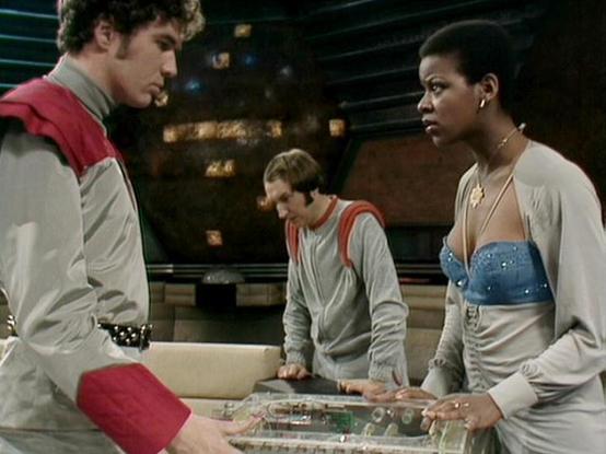 Claude 3.7 describes the image as: "This image appears to be from a science fiction television series, showing a scene set in what looks like a spacecraft or control room. Three people are gathered around what appears to be a transparent tactical table or control console. 

The individuals are wearing distinctive costume styling typical of classic sci-fi productions, with the person on the left wearing a gray and red uniform with shoulder details. The person in the middle wears a simple gray ou…