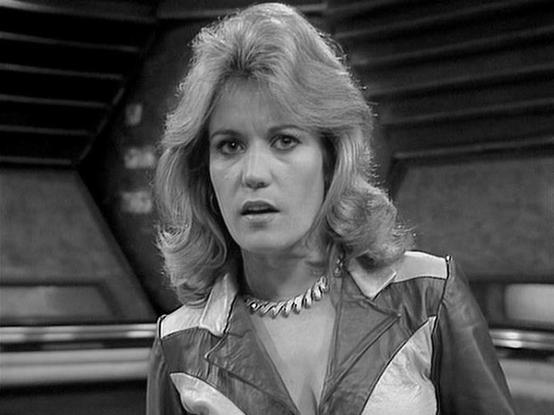 Claude Sonnet 4.0 describes the image as: "This black and white image shows a scene from what appears to be a science fiction television series, likely from the late 1970s or early 1980s based on the styling and production quality. The setting appears to be inside a spacecraft or futuristic facility, with characteristic curved walls and technical details visible in the background that are typical of sci-fi productions from that era.

The scene captures a dramatic moment, with the lighting and c…