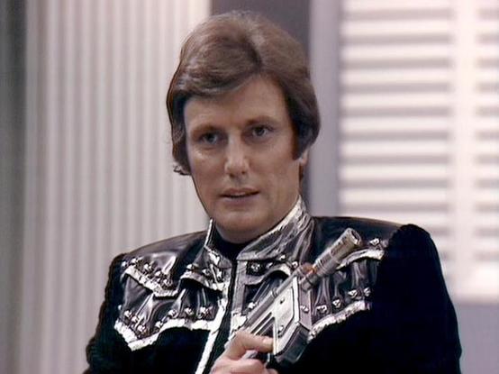 Claude Sonnet 4.0 describes the image as: "This image shows a character in a distinctive black leather outfit with ornate silver studs and decorative metalwork across the chest and shoulders. The costume has a futuristic, militaristic design typical of science fiction television from this era. The character appears to be in an interior setting with white columns and venetian blinds visible in the background, suggesting a formal or institutional environment. The lighting and image quality indica…