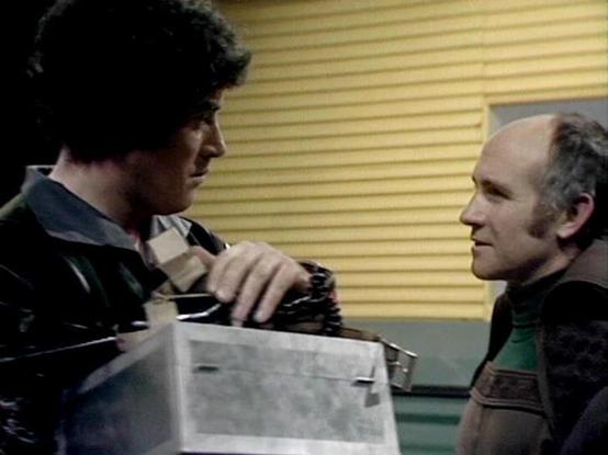 Claude 3.7 describes the image as: "The image shows two individuals in what appears to be a scene from a vintage science fiction television series from the late 1970s or early 1980s. The setting has the characteristic look of a spaceship or futuristic facility with yellow horizontal blinds visible in the background.

One person on the left is holding what looks like a clear box or container, while engaging in conversation with another person on the right who is wearing a dark top with some patt…