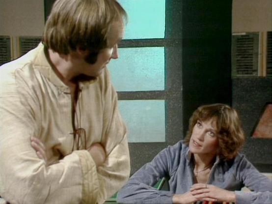 Claude 3.7 describes the image as: "The image shows a scene in what appears to be a futuristic interior setting with distinct architectural elements and lighting visible in the background. 

On the left is a person with short hair wearing a light-colored, loose-fitting garment with ties at the front, standing with arms crossed. On the right is someone with wavy brown hair wearing a blue-gray outfit, seated and looking up at the standing figure.

The composition suggests this is from a science f…