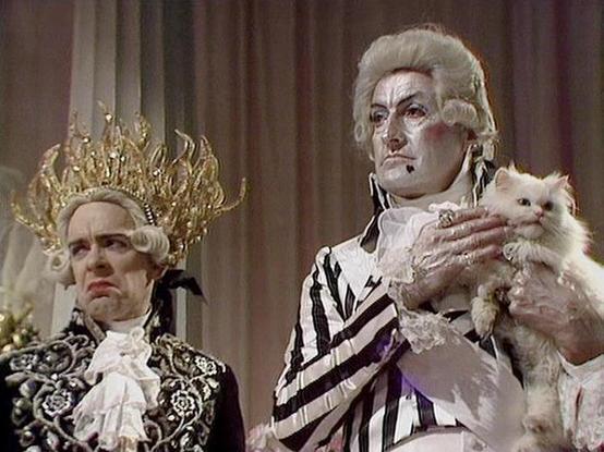 Claude Sonnet 4.0 describes the image as: "I can see this is a scene featuring two characters in elaborate period costumes with powdered wigs, suggesting an 18th-century aristocratic setting. One character wears ornate clothing with decorative embroidery and an elaborate golden flame-like crown or headdress. The other character is dressed in a striped robe or dressing gown and is holding what appears to be a small white cat or similar pet. The setting appears formal and palatial, with classical…