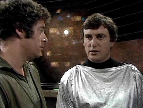 Claude Sonnet 4.0 describes the image as: "I can see two men in what appears to be a futuristic or sci-fi setting with metallic walls and atmospheric lighting in the background. One man is wearing a dark green or olive-colored shirt, while the other is dressed in a distinctive silver metallic garment with a black collar or turtleneck underneath. The scene appears to be taking place in some kind of spaceship or advanced facility interior. The lighting and set design have the characteristic look …