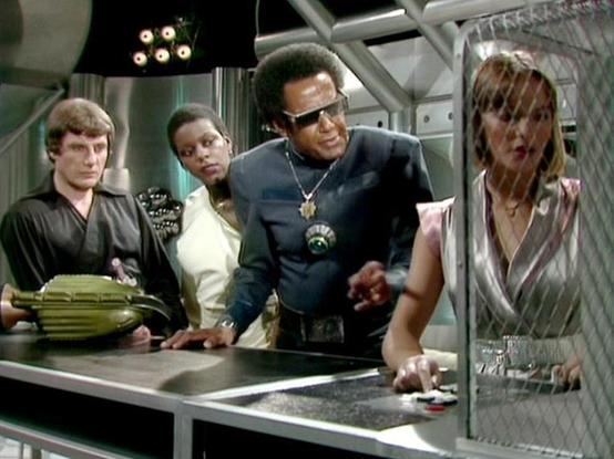 Claude Sonnet 4.0 describes the image as: "I can see this appears to be from a science fiction television series, showing what looks like a futuristic spacecraft interior or control room setting. The scene features several people gathered around what appears to be a control console or table with some kind of device on it. The set design has that distinctive late 1970s/early 1980s sci-fi aesthetic with white and metallic surfaces, and there are some lighting effects visible in the background. Th…