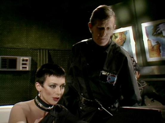 Claude 3.7 describes the image as: "The image shows a dark, futuristic interior setting with a distinctly retro sci-fi aesthetic typical of British television productions from the late 1970s/early 1980s. 

In the foreground is a person with short dark hair wearing a distinctive high collar or choker and elegant attire. Behind them stands another figure in a dark uniform or outfit that appears military or authoritarian in style.

The scene appears to be taking place in some kind of control room …