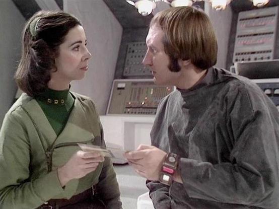 Claude 3.7 describes the image as: "This image shows a scene in what appears to be a futuristic spacecraft or control room setting. Two people are engaged in conversation, with one wearing a green outfit with a light green overlay jacket, and the other in a gray outfit with what looks like a wrist device or communication tool. They appear to be exchanging some kind of document or data card.

The setting features technological elements typical of science fiction production design from the late 1…