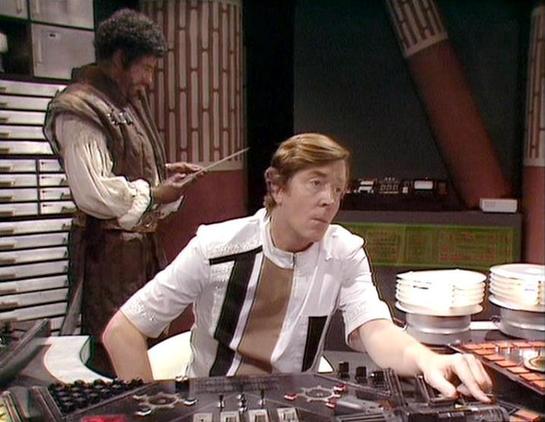Claude Sonnet 4.0 describes the image as: "I can see this is a scene set on what appears to be a spaceship bridge or control room, with characteristic sci-fi production design featuring control panels, lighting, and futuristic set pieces typical of late 1970s British television. The setting shows sophisticated electronic equipment and displays. One person is seated at what looks like a control console or computer terminal, wearing a light-colored outfit with distinctive black trim details. Anot…