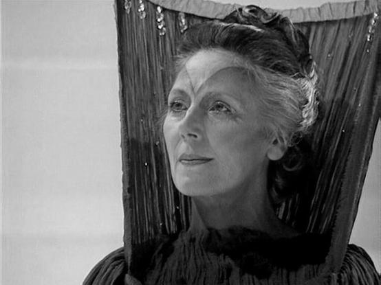 Claude 3.7 describes the image as: "This is a black and white still from a television production, showing a person in what appears to be period costume with their hair styled in an elegant updo. The image has a dramatic quality with the subject framed by what looks like decorative curtains or textured backdrop material with some subtle shimmering elements.

The composition creates a theatrical feel, with the lighting creating strong contrasts typical of classic film or television drama. The sty…