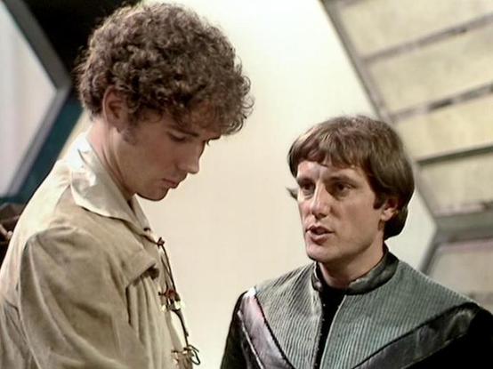 Claude 3.7 describes the image as: "The image shows a scene from a science fiction television production with a futuristic setting. Two individuals are captured in conversation, with one wearing a light-colored tunic with decorative fastenings, and the other in a dark green or gray high-collared outfit that appears to be made of a textured material. The person on the left has curly hair and is looking down, while the person on the right has shorter, straight hair and appears to be speaking. The…