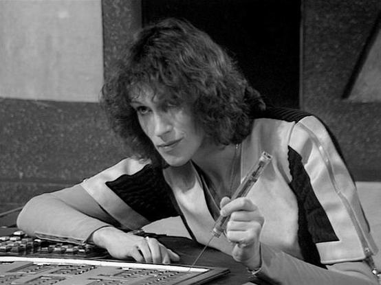 Claude Sonnet 4.0 describes the image as: "I can see this is a black and white photograph showing someone working at what appears to be a control console or technical equipment. The person has curly hair and is wearing what looks like a light-colored top or vest. They appear to be focused on operating or examining some kind of electronic device or control panel, holding what might be a tool or instrument. The setting appears to be inside some kind of technical or futuristic environment, consist…