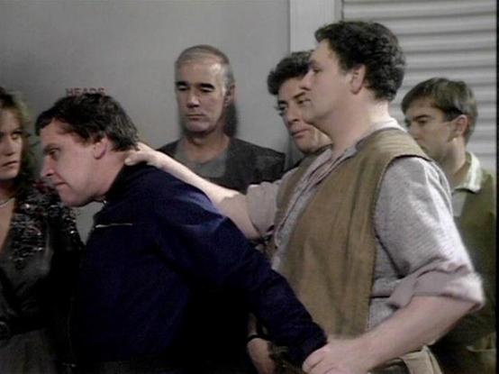 Claude 3.7 describes the image as: "The image shows a scene from what appears to be a sci-fi television program from the late 1970s or early 1980s, based on the production quality and styling. The scene depicts a tense moment with several people in what looks like a corridor or interior room with gray walls.

In the foreground, there's an interaction where one person in a light-colored vest is restraining or holding someone in a dark blue outfit. Several others stand in the background observing…