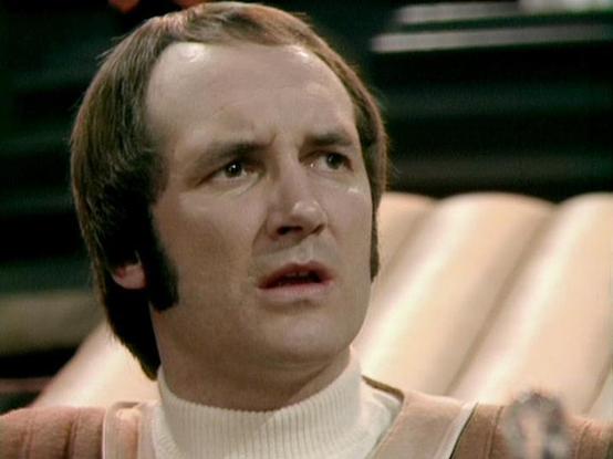Claude 3.7 describes the image as: "The image shows a person with a concerned or surprised expression in what appears to be a scene from a vintage television production, likely from the late 1970s or early 1980s based on the filming style and aesthetic. 

The individual is wearing a light-colored turtleneck sweater and has distinctive sideburns and a hairstyle characteristic of that era. The background appears to be part of a set with some green elements visible.

The lighting and film quality …