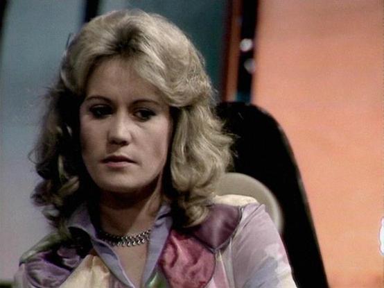 Claude 3.7 describes the image as: "The image appears to be from a vintage television program, likely from the late 1970s or early 1980s based on the production style and aesthetic. It shows a person with blonde, voluminous feathered hair typical of that era. They are wearing a colorful outfit with pastel pink and purple tones and a metallic choker necklace. The background suggests a studio set with soft lighting and muted colors common in science fiction productions of that period. The scene h…