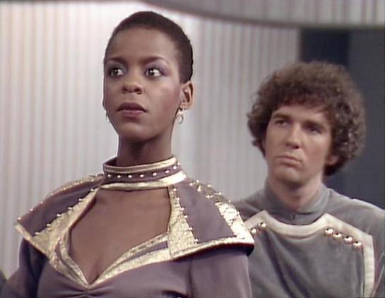 Claude 3.7 describes the image as: "The image shows a scene from what appears to be a science fiction television series from the late 1970s or early 1980s, based on the production style and costumes. 

Two performers are shown in futuristic attire featuring distinctive metallic accents and structured shoulders typical of space-themed productions of that era. The foreground figure wears a purple/gray outfit with gold trim and decorative elements around the collar, while the person in the backgro…