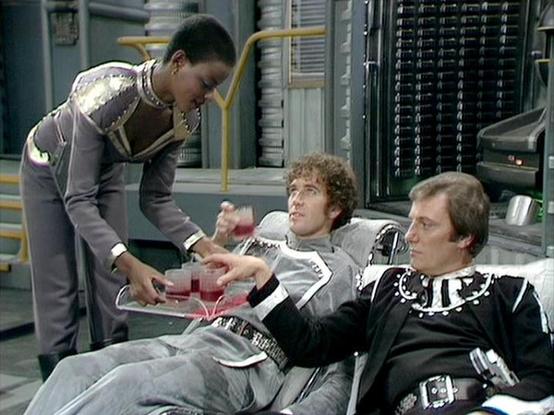 Claude Sonnet 4.0 describes the image as: "This scene appears to be set in a futuristic medical bay or treatment facility aboard a spacecraft. The sterile, high-tech environment features metallic walls and sophisticated equipment typical of science fiction settings. Two individuals are seated in what appear to be medical treatment chairs, while a third person in a form-fitting uniform is attending to them, holding what looks like medical equipment or monitoring devices. The setting suggests thi…
