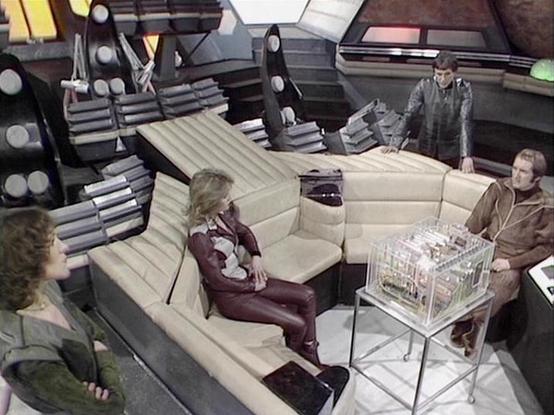 Claude Sonnet 4.0 describes the image as: "This appears to be a scene set in the crew quarters or common area of a spacecraft. The setting features a curved, cream-colored seating area with a futuristic design typical of science fiction television from this era. The interior has a utilitarian, spaceship aesthetic with visible structural elements and control panels in the background.

Several crew members are present in the scene, positioned around the seating area. The staging suggests this is …